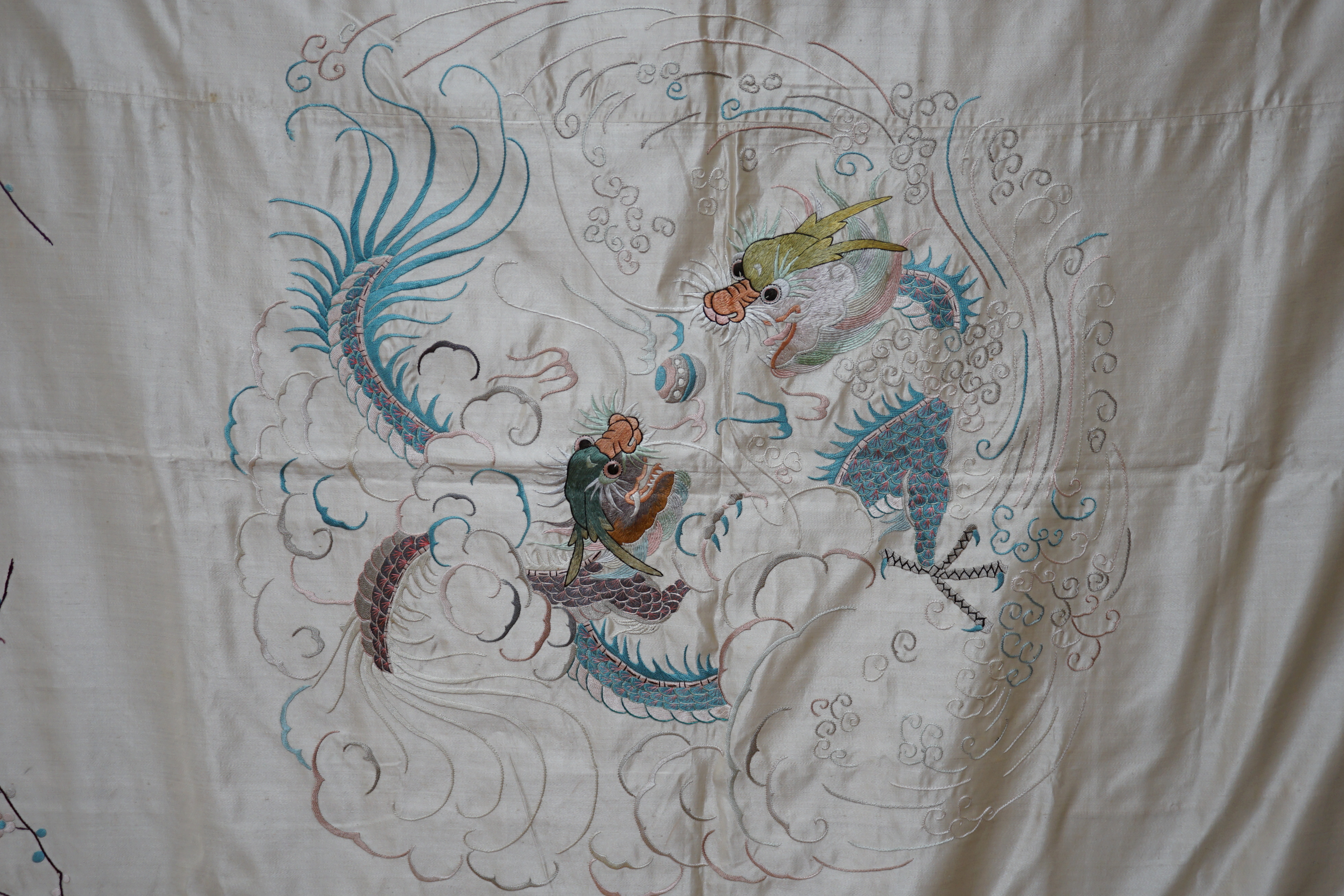 A 1920’s-30’s Chinese oyster silk satin embroidered shawl, now converted to a wall hanging, embroidered in pastel coloured silk threads, the large central motif of two dragons playing with a pearl, the corners of the han